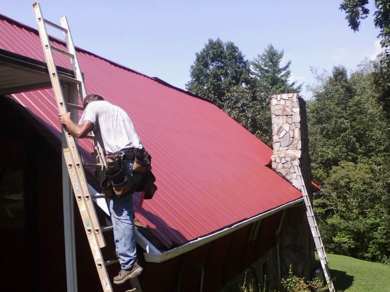 Roofer on a ladder beside a red metal roof and stone chimney — Soffit & Fascia Repair by Roofing Brothers in New Trenton